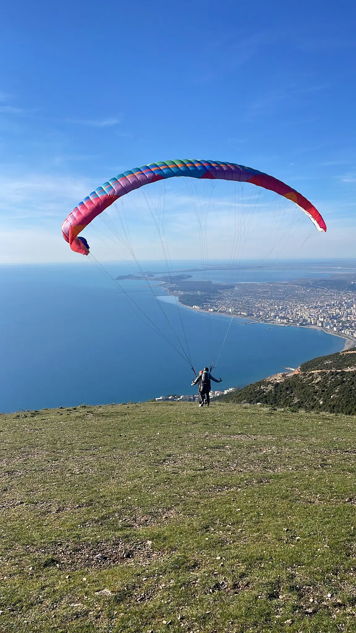 paraglider launching from a grassy hill overlooking a coastal city
